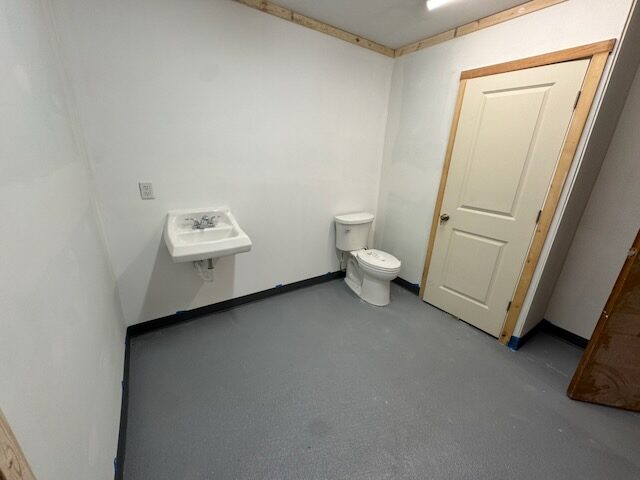 Bathroom with a wall-mounted sink, toilet, finished drywall, and a gray floor, with the door at right
