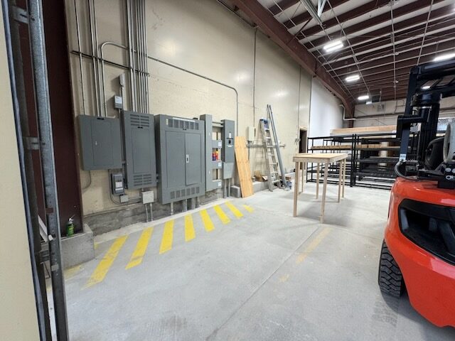 Electrical panels and conduit on the shop wall beside a yellow striped floor marking, with racking and a forklift in the background