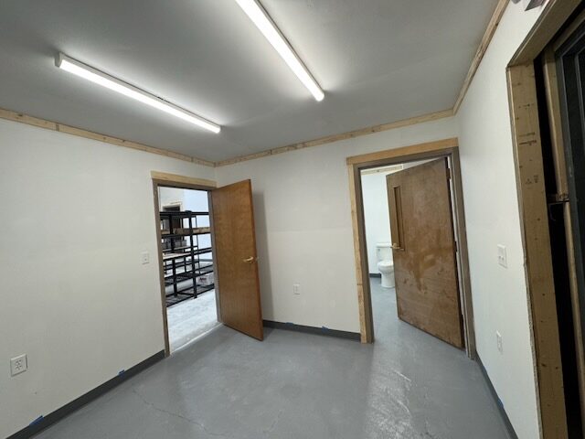 Office with finished drywall and a gray floor, with an open door to the shop area and racking and a second door to a bathroom
