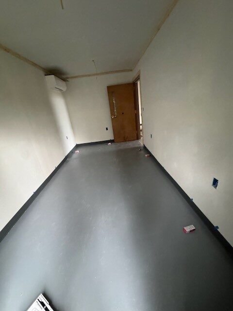 Long narrow room with a wall-mounted mini split, painted drywall, dark base trim, and a gray floor, looking toward a wood door